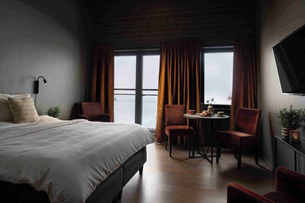 A hotel room with a large bed, a desk with a chair, a small table with a plant, and a TV on the wall.