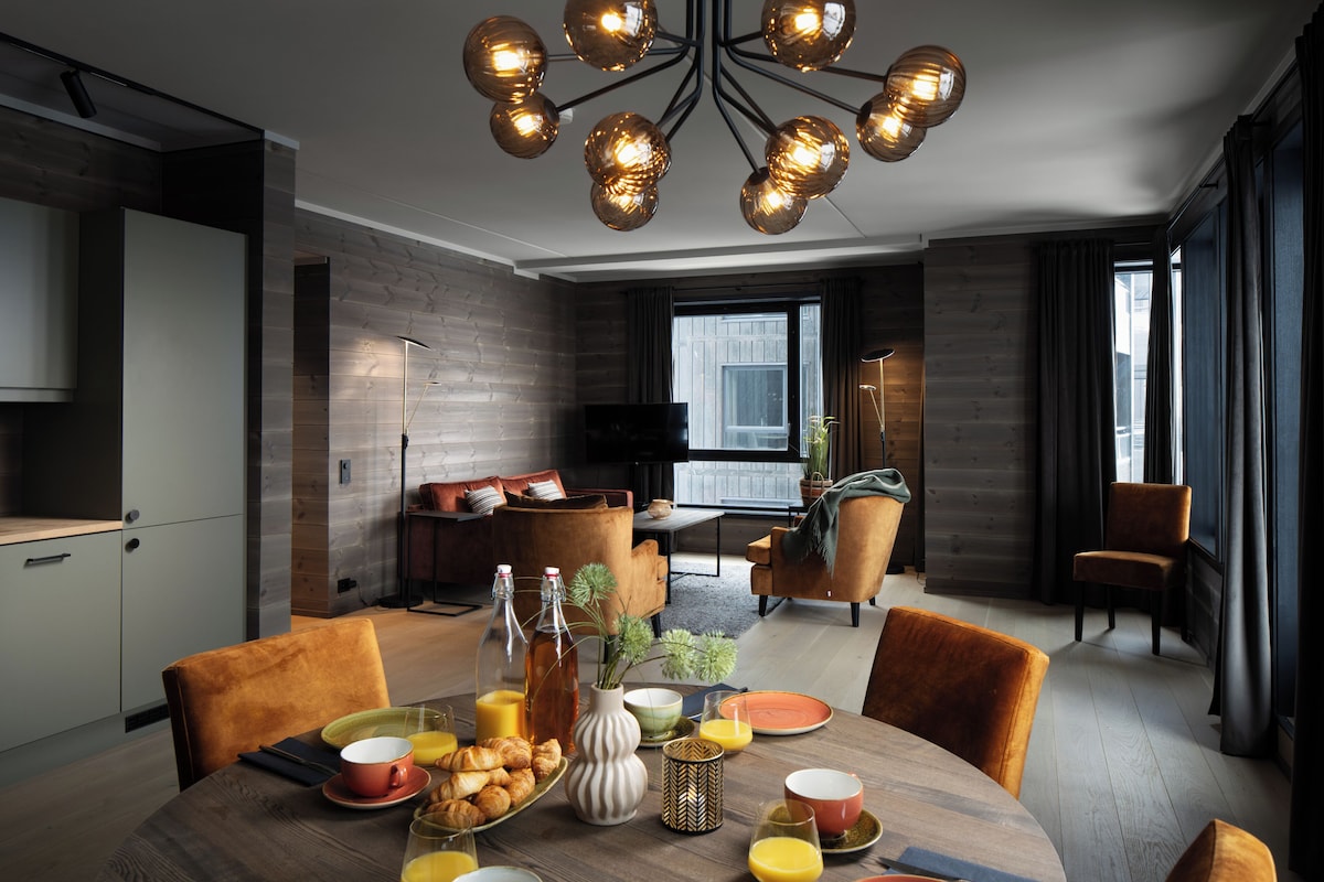 A modern dining area with a wooden table set for breakfast, a chandelier, and a view of the city through large windows.