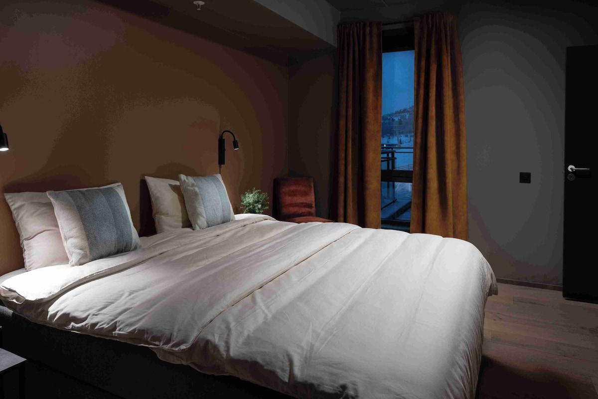 A neatly made bed with white linens and multiple pillows, a bedside lamp, and a view of a balcony through a window with curtains.