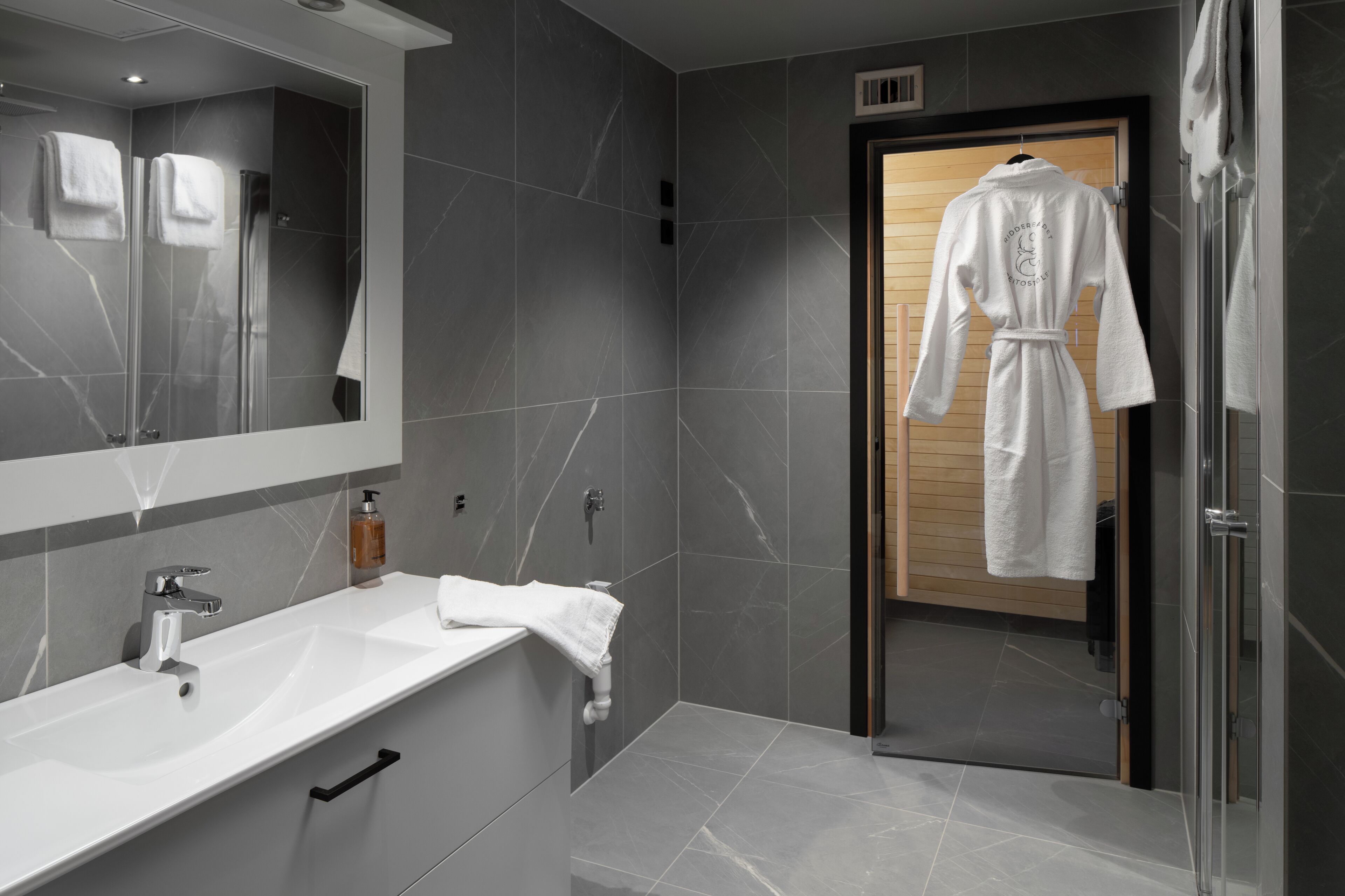 A modern bathroom with a large mirror, a white sink, and a robe hanging on a door.