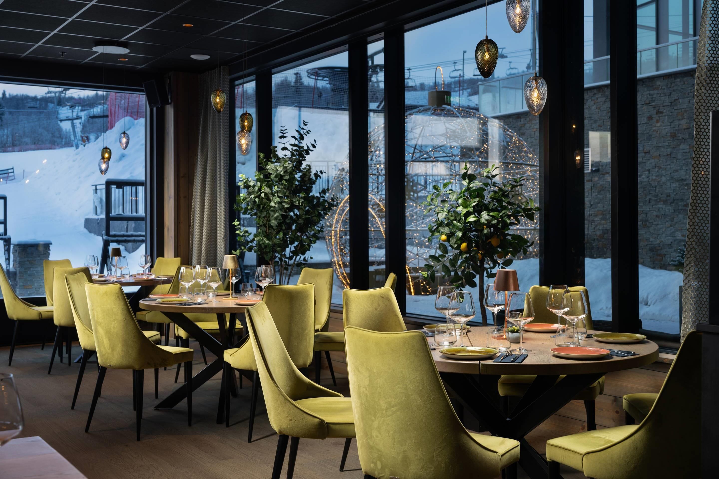 Un restaurante con sillas amarillas, mesas redondas y grandes ventanales con vistas a un paisaje nevado.