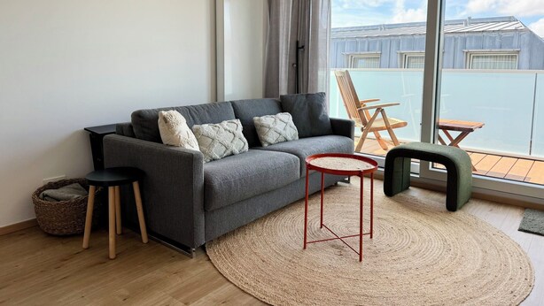 A modern living room with a grey sofa, a small round table, and a view of a balcony with outdoor furniture. - Kampstraße; Wohnung 20 (Norderney)