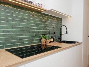 Une cuisine avec un mur recouvert de carreaux verts, un plan de travail en bois, un robinet noir et une étagère où sont posées des tasses colorées.