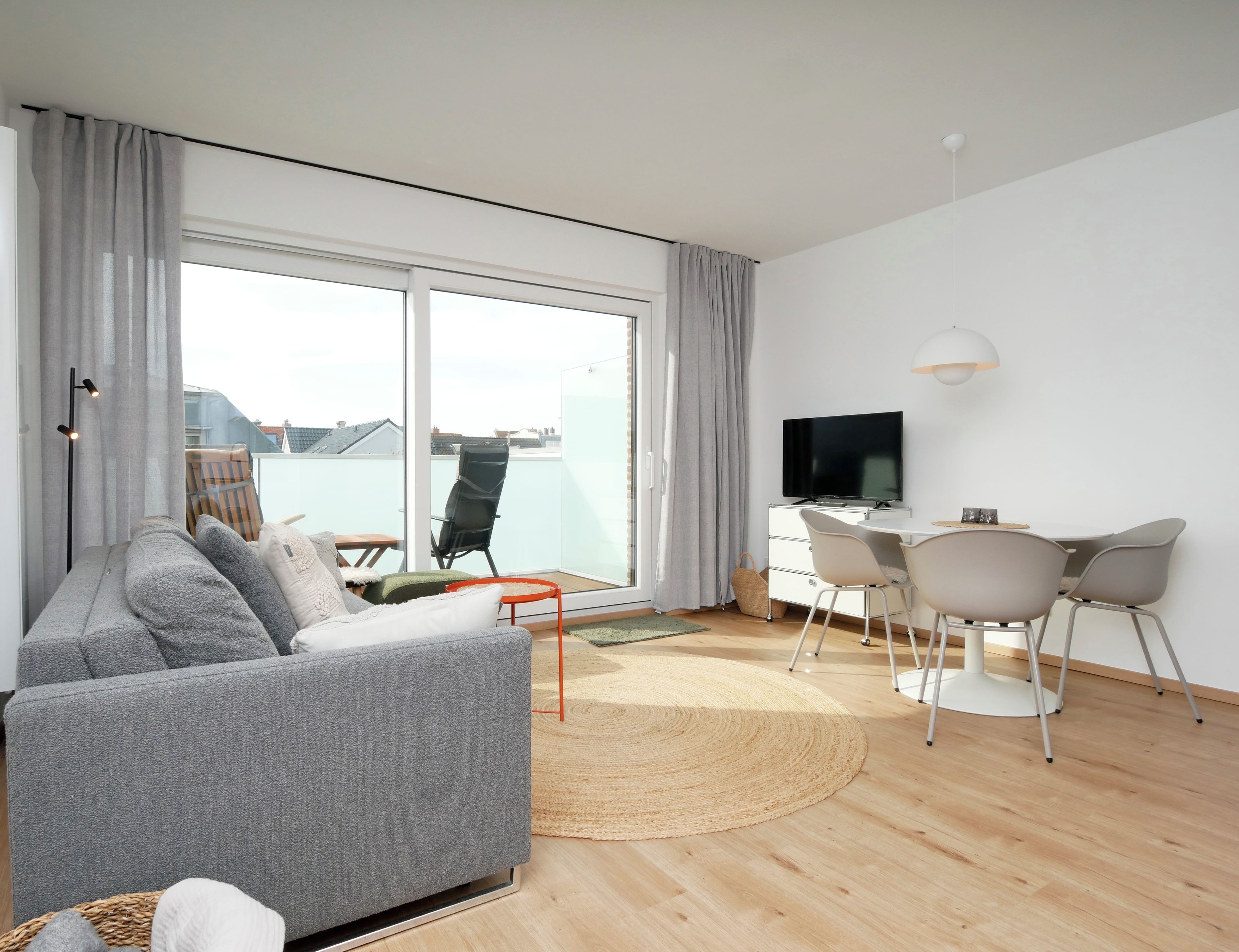 A modern living room with a sofa, a dining table with chairs, a flat-screen TV, and a large window with curtains.