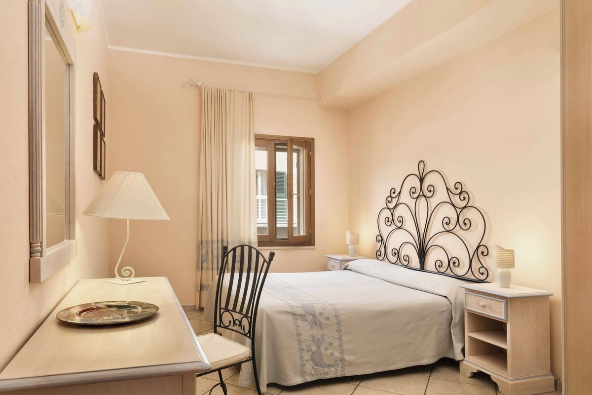 A bedroom with a bed, a desk, a chair, a nightstand, and a window with curtains.