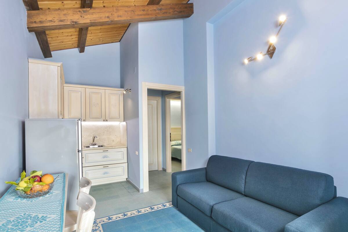 A compact living space with a kitchenette, a sofa, and a dining table with fruit.