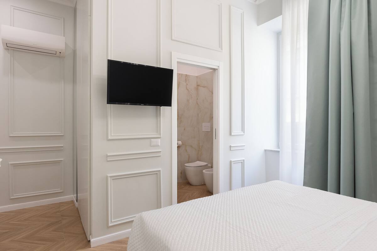 A hotel room with a bed, a television, a bathroom with a toilet and sink, and a door leading to another room.