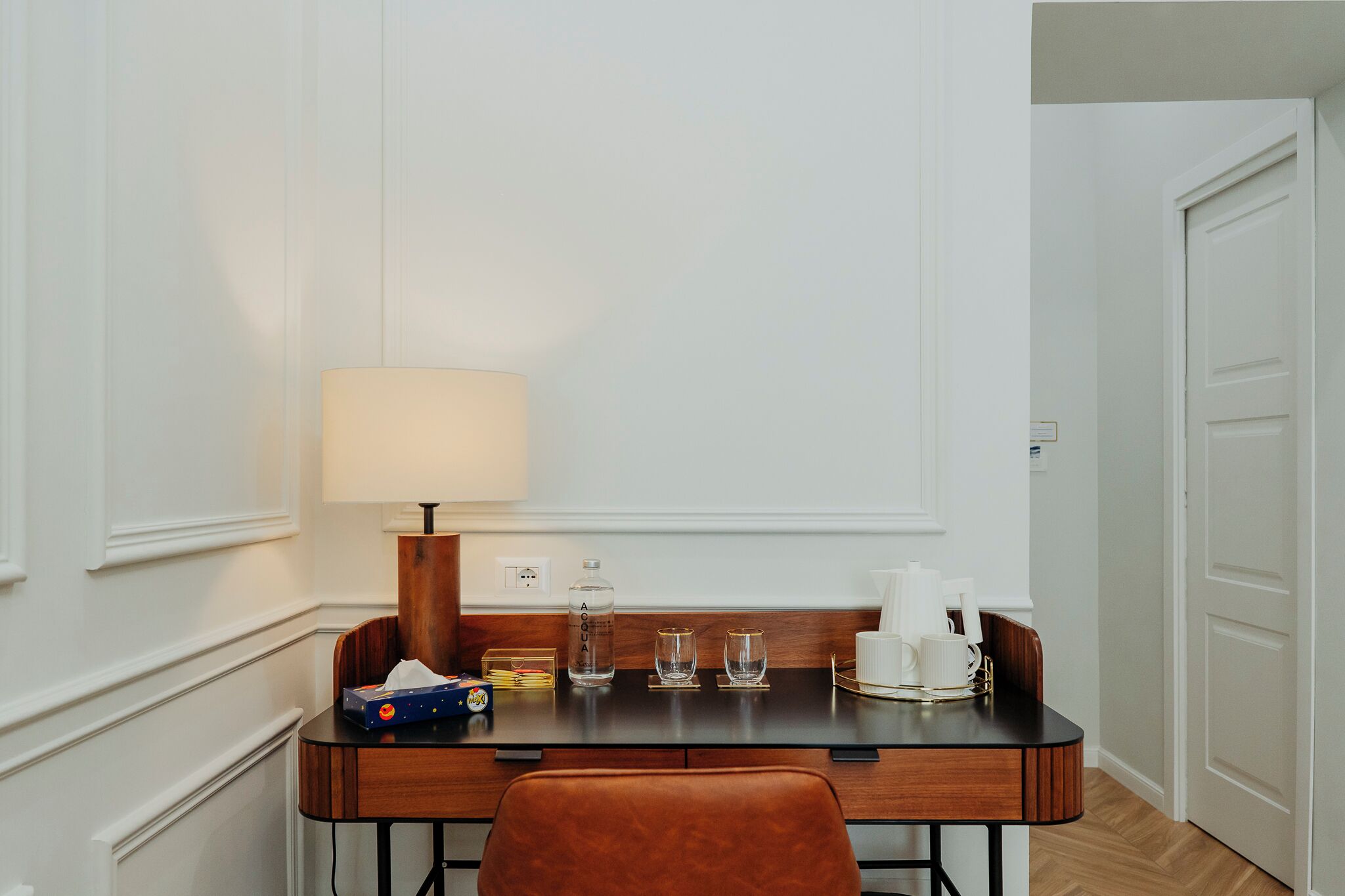 A hotel room with a wooden desk, a lamp, a bottle of water, two glasses, a tissue box, and a coffee maker.