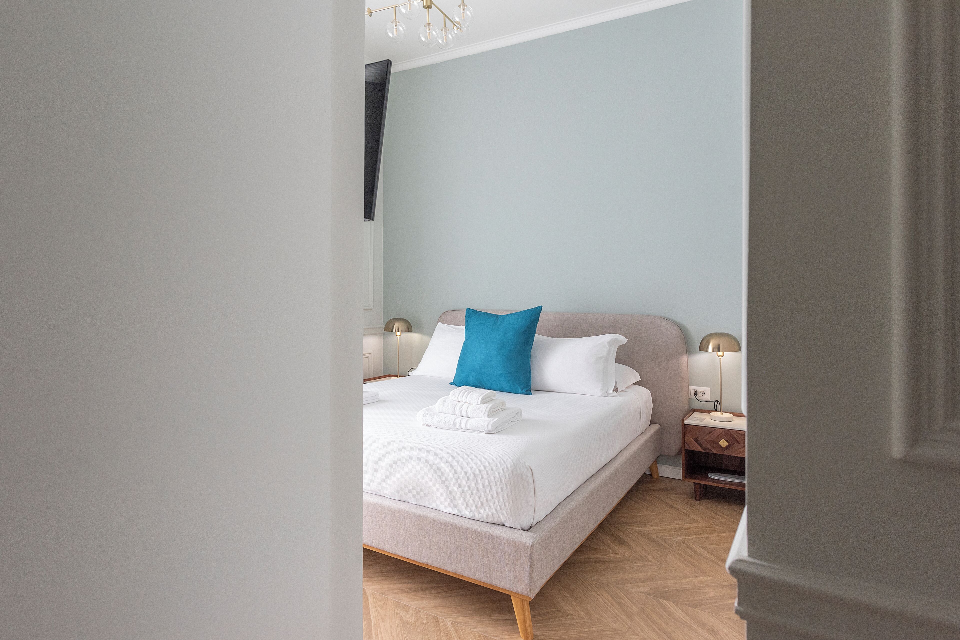 A neatly made bed with white linens and a blue pillow, a bedside table with a lamp, and a wooden floor.