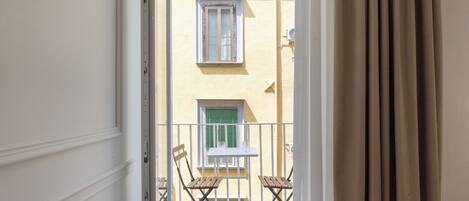 A balcony with two wooden chairs and a small table, overlooking a building with a yellow facade and green shutters.
