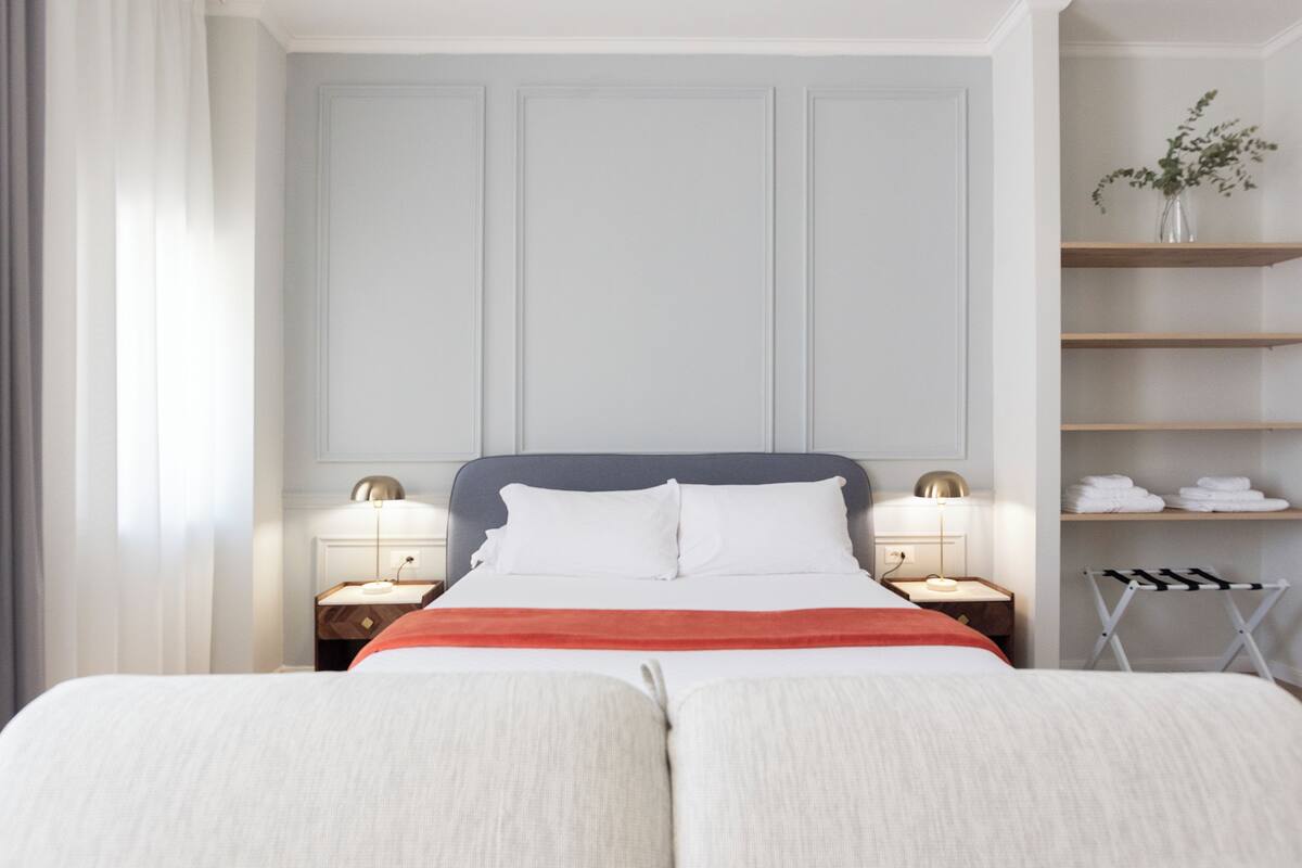 A hotel room with a large bed, two bedside tables, a nightstand, a shelf with towels, and a window with curtains.