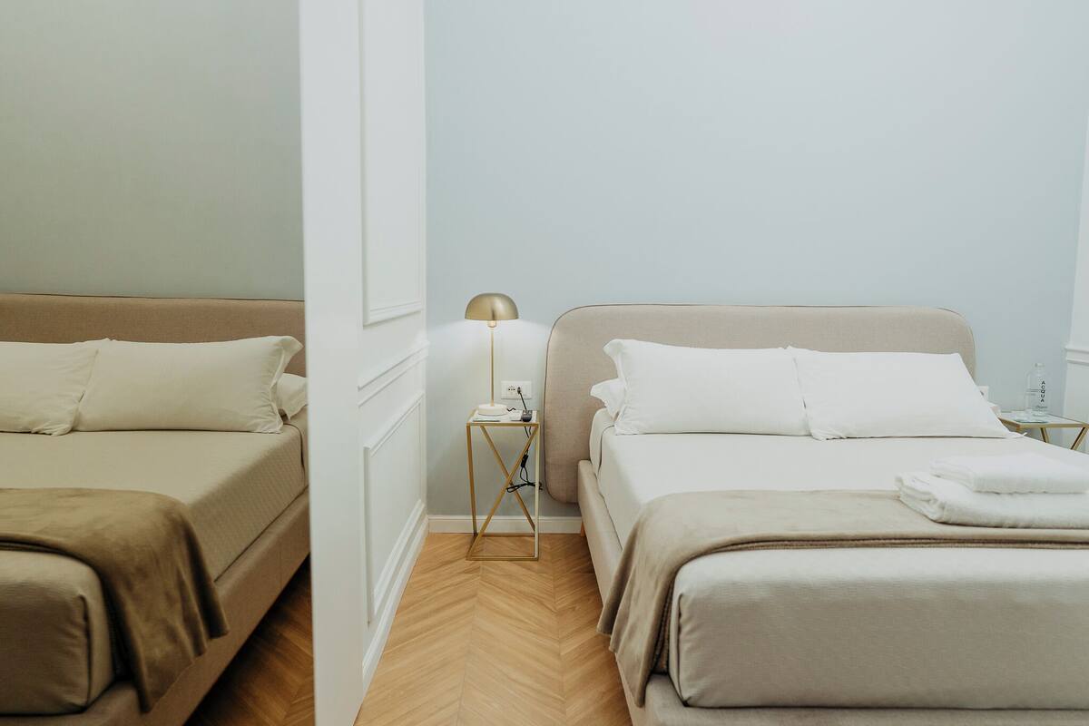 A hotel room with two beds, a bedside table with a lamp, and a herringbone-patterned wooden floor.