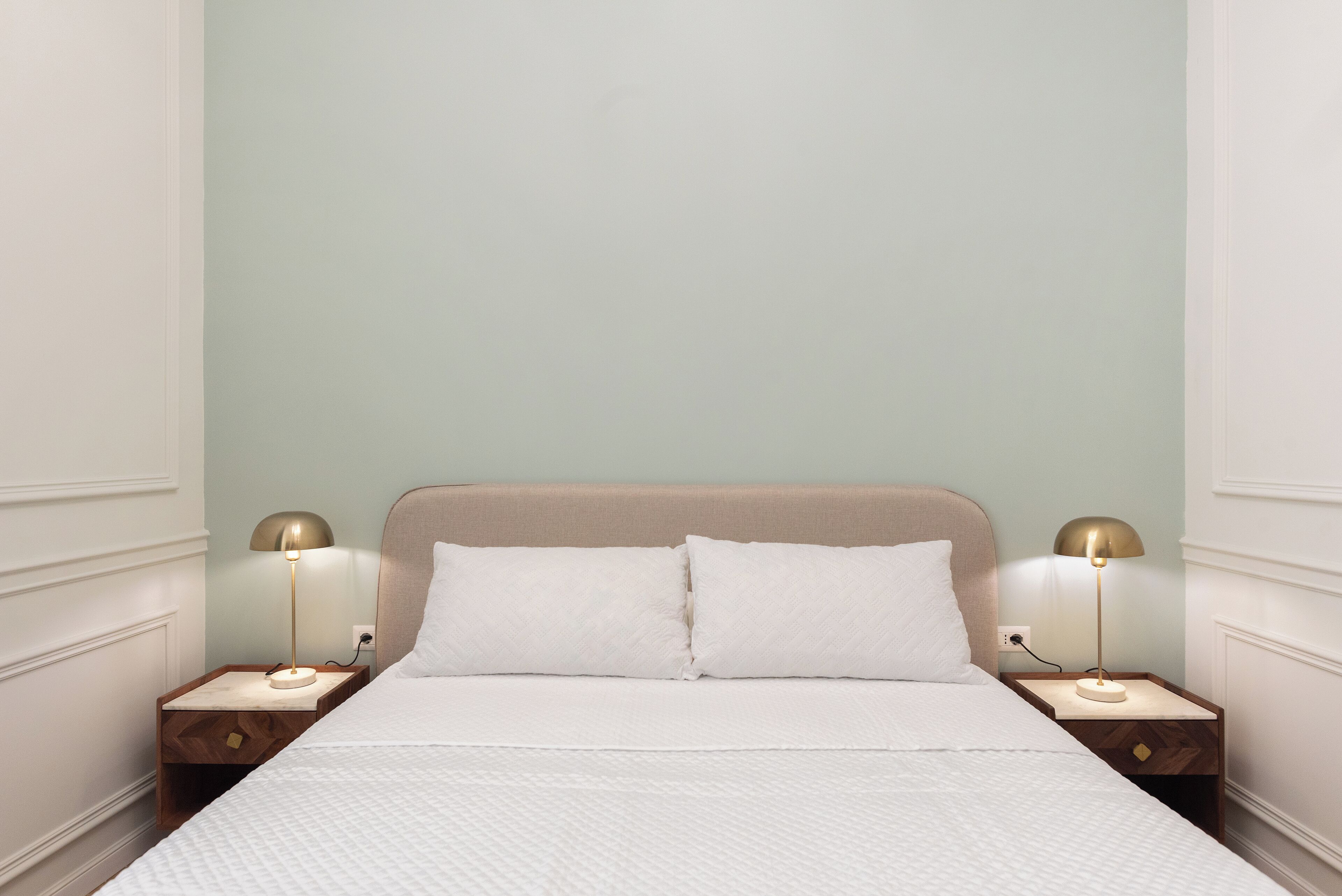 Una cama bien hecha con sábanas blancas, dos mesitas de noche con lámparas y una pared de color verde claro.