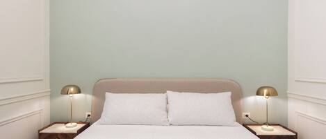Una cama bien hecha con sábanas blancas, dos mesitas de noche con lámparas y una pared de color verde claro.