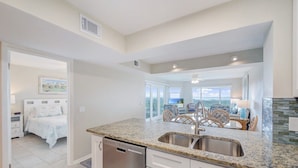 A modern kitchen with granite countertops, a dishwasher, and a dining area with wicker chairs. - Colony Reef Club 1203 - 3 Bedroom Oceanfront, 2nd floor with elevator access (St. Augustine Beach)