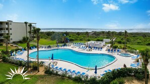 A large, curved swimming pool with blue lounge chairs and umbrellas, surrounded by greenery and a view of the ocean. - Colony Reef Club 1203 - 3 Bedroom Oceanfront, 2nd floor with elevator access (St. Augustine Beach)