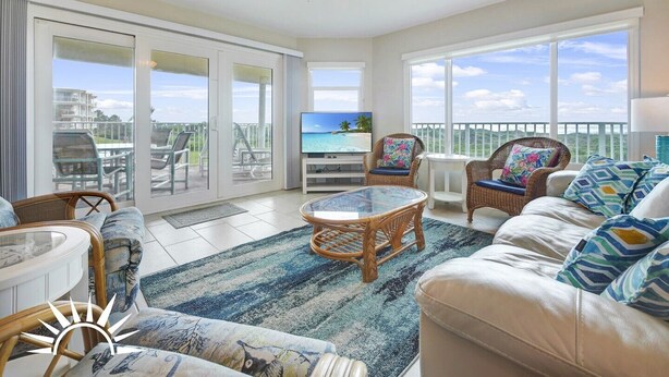 A spacious living room with a ceiling fan, a television, wicker furniture, and large windows offering a view of the beach. - Colony Reef Club 1203 - 3 Bedroom Oceanfront, 2nd floor with elevator access (St. Augustine Beach)