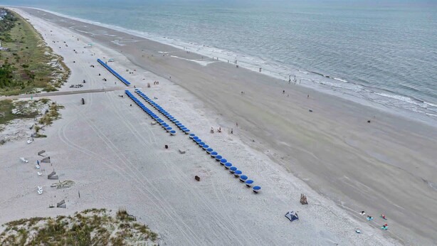 Ein Strand mit blauen Sonnenschirmen und Liegestühlen, ein sandiger Uferstreifen und das Meer im Hintergrund.