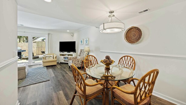 A dining area with a glass table and wicker chairs, a ceiling light, and a television in the background. - Fresh & Modern Villa by the Sea Beach Club Getaway (Hilton Head Island)