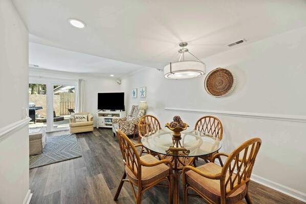 A dining area with a glass table and wicker chairs, a ceiling light, and a television in the background. - Fresh & Modern Villa by the Sea Beach Club Getaway (Hilton Head Island)