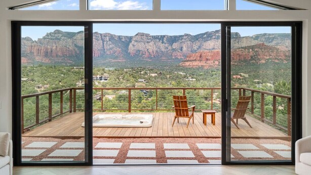 A spacious balcony with a wooden deck, two wooden chairs, and a glass door leading to a mountain view. - Best of Sedona Views, Cliffside Deck, Fitness/Yoga Room and More! (Sedona)