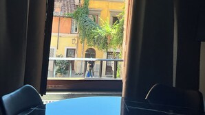 Uitzicht vanuit een raam met gordijnen, op een geel gebouw met meerdere ramen en balkons, en een boom op de voorgrond.