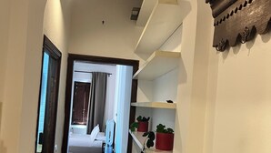 Een hal met een betegelde vloer, witte muren en een houten plafond. Er staan planken met potplanten en een deur naar een andere kamer.