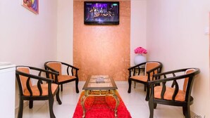 Una habitación con televisión montada en la pared, una mesa de vidrio y un tapete rojo, además de cuatro sillas con cojines naranjas.