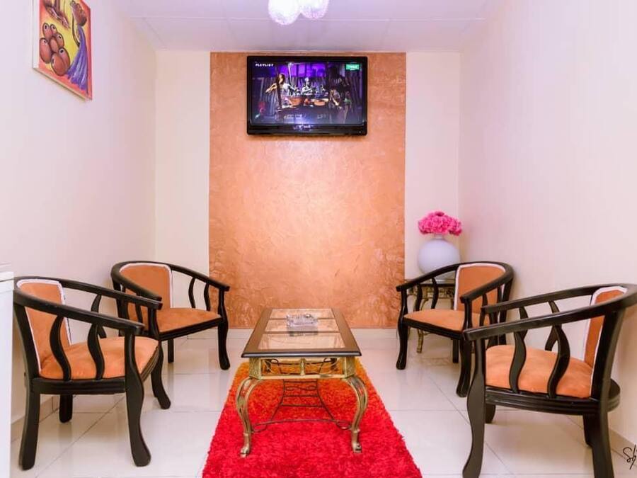 Una habitación con televisión montada en la pared, una mesa de vidrio y un tapete rojo, además de cuatro sillas con cojines naranjas.