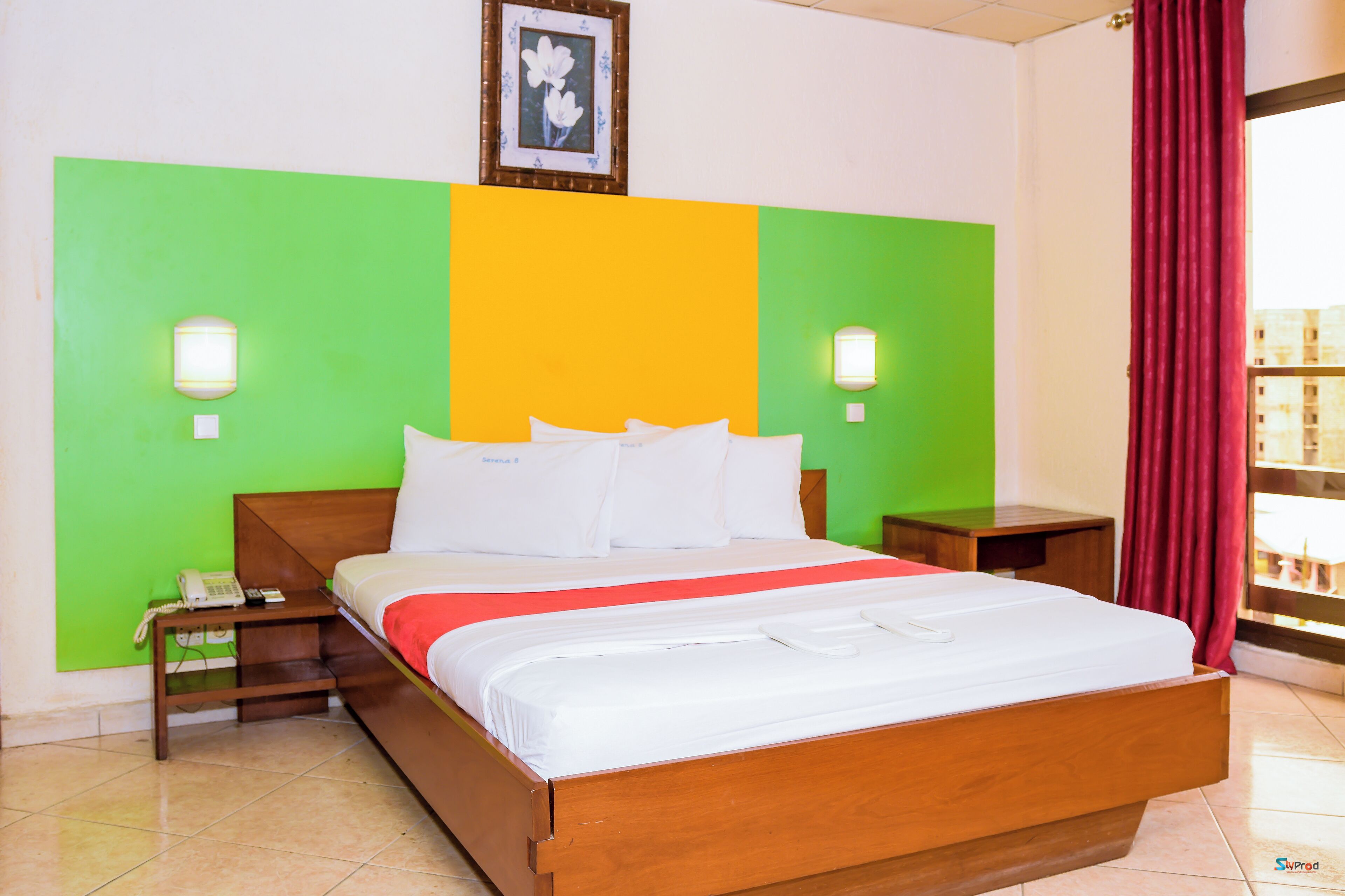 Habitación de hotel con cama de madera, paredes decoradas en tonos verde y amarillo, cortina roja y un cuadro en la pared.