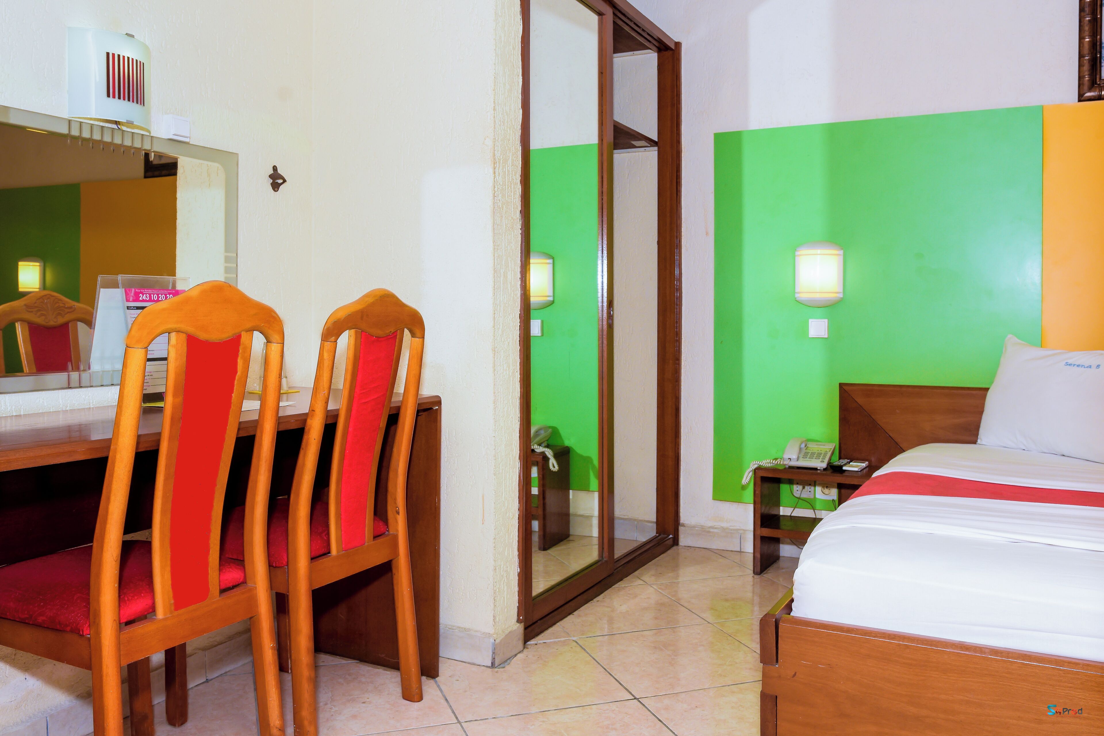 A hotel room with a bed, a desk with a chair, a mirror, a nightstand with a phone, and a green wall.