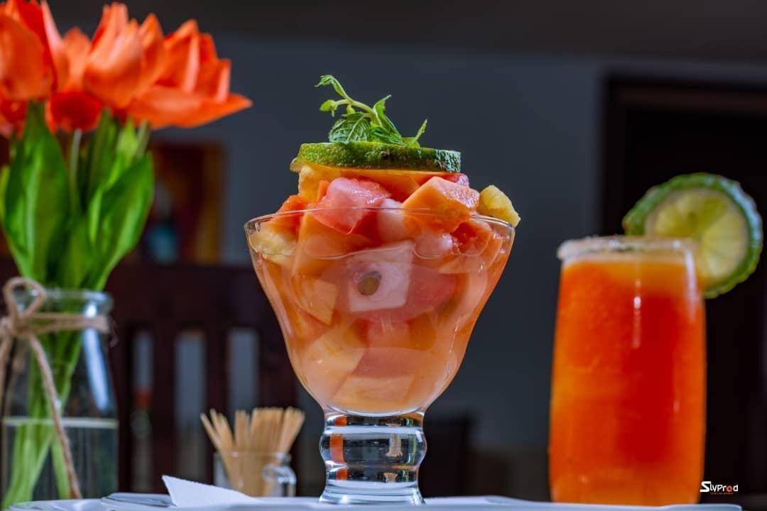 A fruit salad in a glass bowl with a garnish, a glass of red drink, and a vase of orange flowers.
