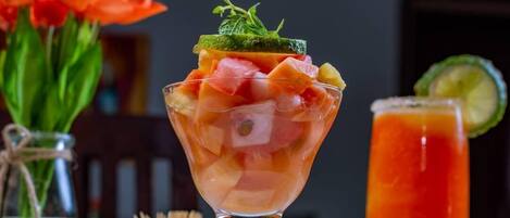 A fruit salad in a glass bowl with a garnish, a glass of red drink, and a vase of orange flowers.