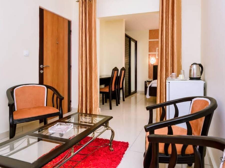 A compact living space with a glass table, two chairs, a red rug, and a kitchenette area.
