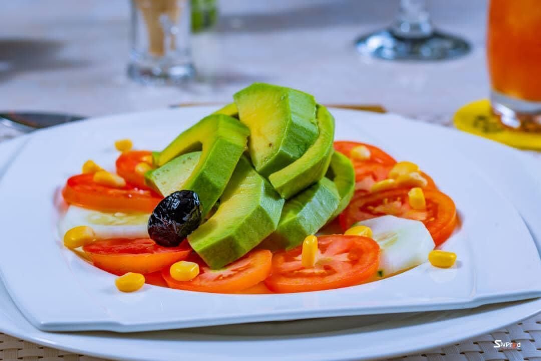 A plate of sliced avocado, tomatoes, corn, and a black olive.