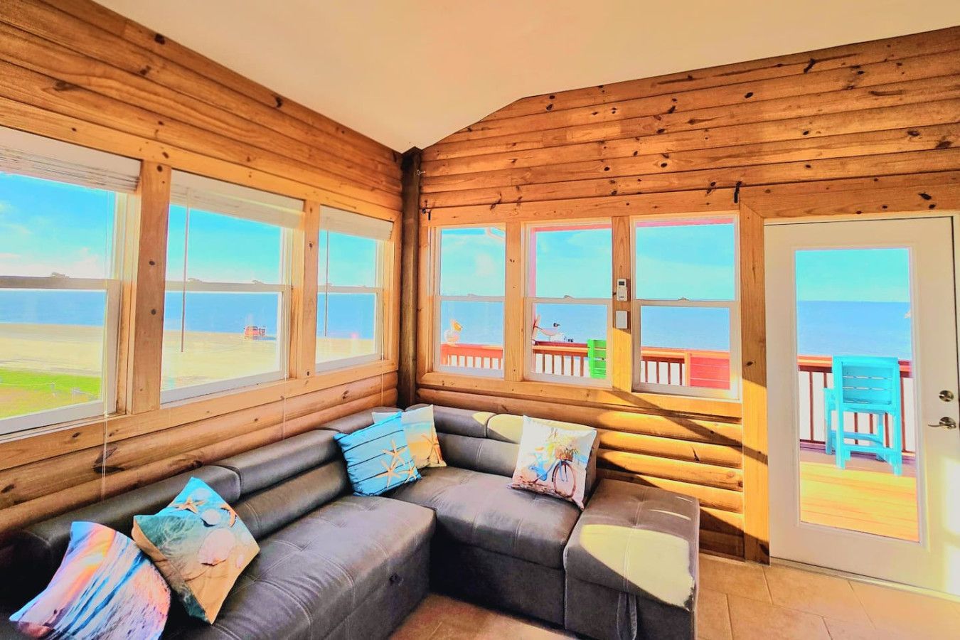 Une chambre aménagée dans un chalet en rondins, dotée d’une grande fenêtre, d’un canapé d’angle gris agrémenté de coussins décoratifs et offrant une vue sur la plage.