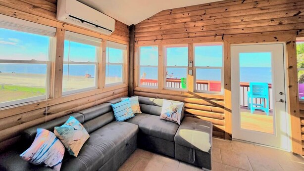 Quarto de cabana de tronco com sofá seccional cinza, janelas amplas e vista para a praia.