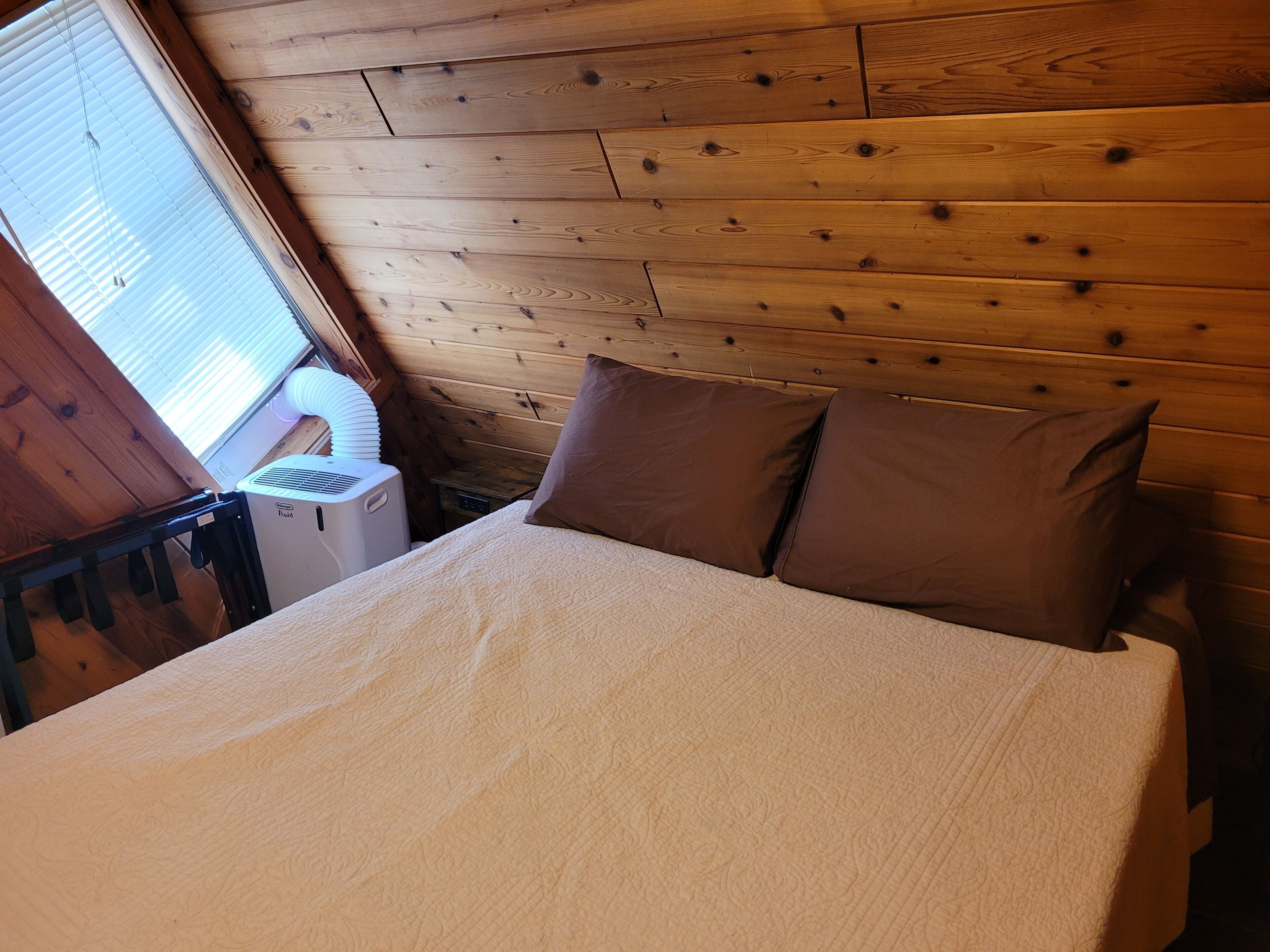 Ein Zimmer in einer Holzhütte mit einem Bett, Kissen, einem Heizgerät und einem Fenster mit Jalousien.