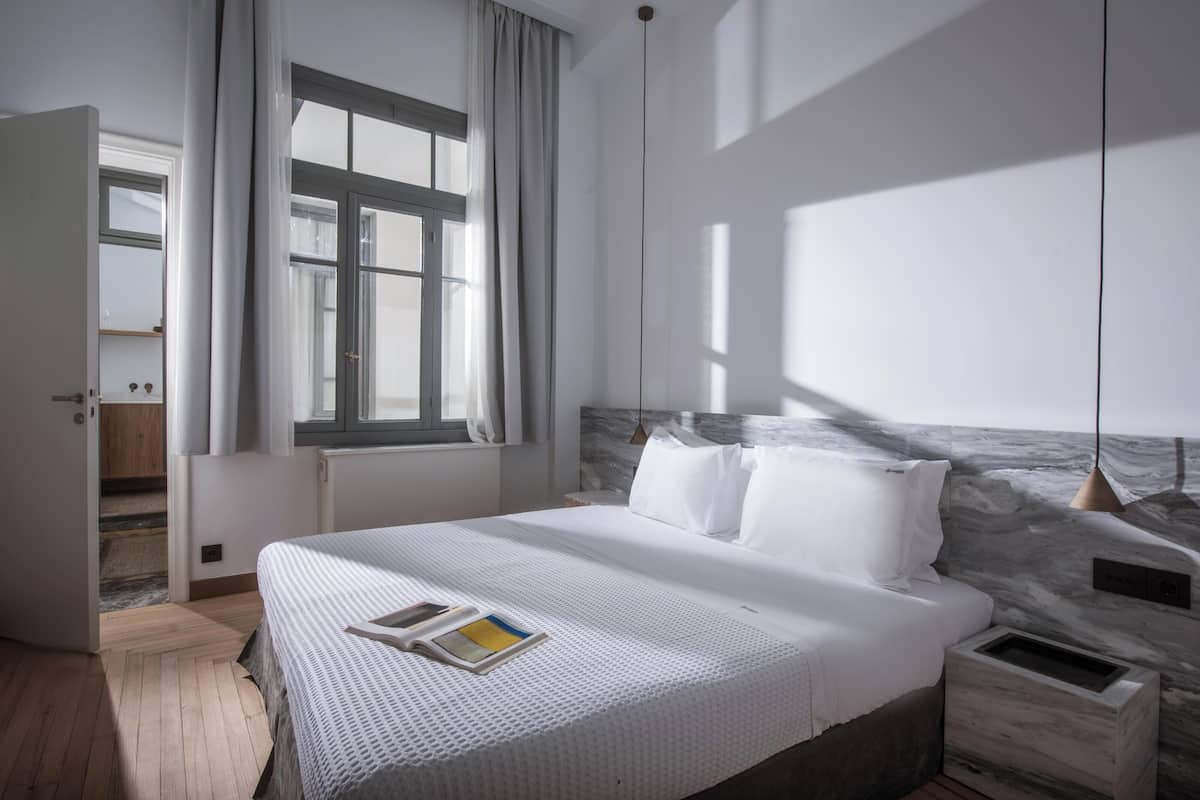 A modern hotel room with a large bed, a marble headboard, a bedside table, and a window with curtains.