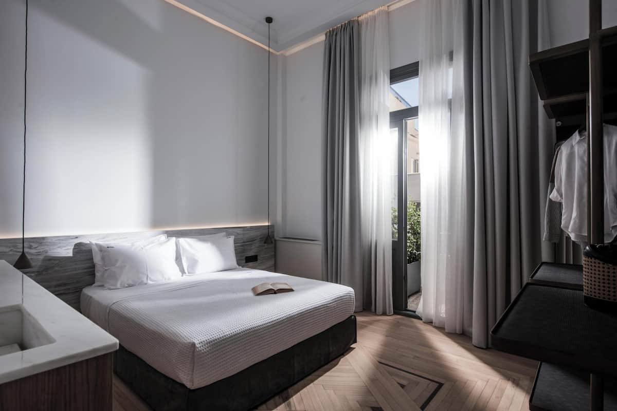 A modern hotel room with a large bed, a bedside table, a built-in wardrobe, and a view through a glass door to the outdoors.