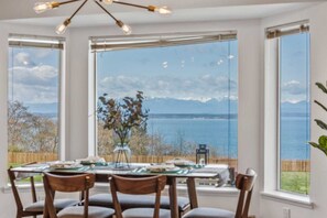 Un coin repas avec une table dressée pour quatre personnes, de grandes fenêtres offrant une vue sur un plan d’eau et des montagnes, et un vase rempli de fleurs séchées posé sur le rebord de la fenêtre.