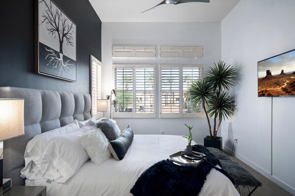 A bedroom with a large bed, a window with blinds, a potted plant, and artwork on the walls.