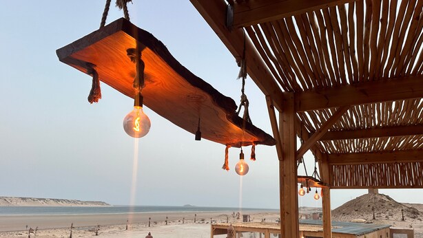 A wooden outdoor seating area with hanging light fixtures, a view of the beach, and a desert landscape in the background. - NORTH LAGOON DAKHLA (Dakhla)