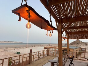 A wooden outdoor seating area with hanging light fixtures, a view of the beach, and a desert landscape in the background. - NORTH LAGOON DAKHLA (Dakhla)