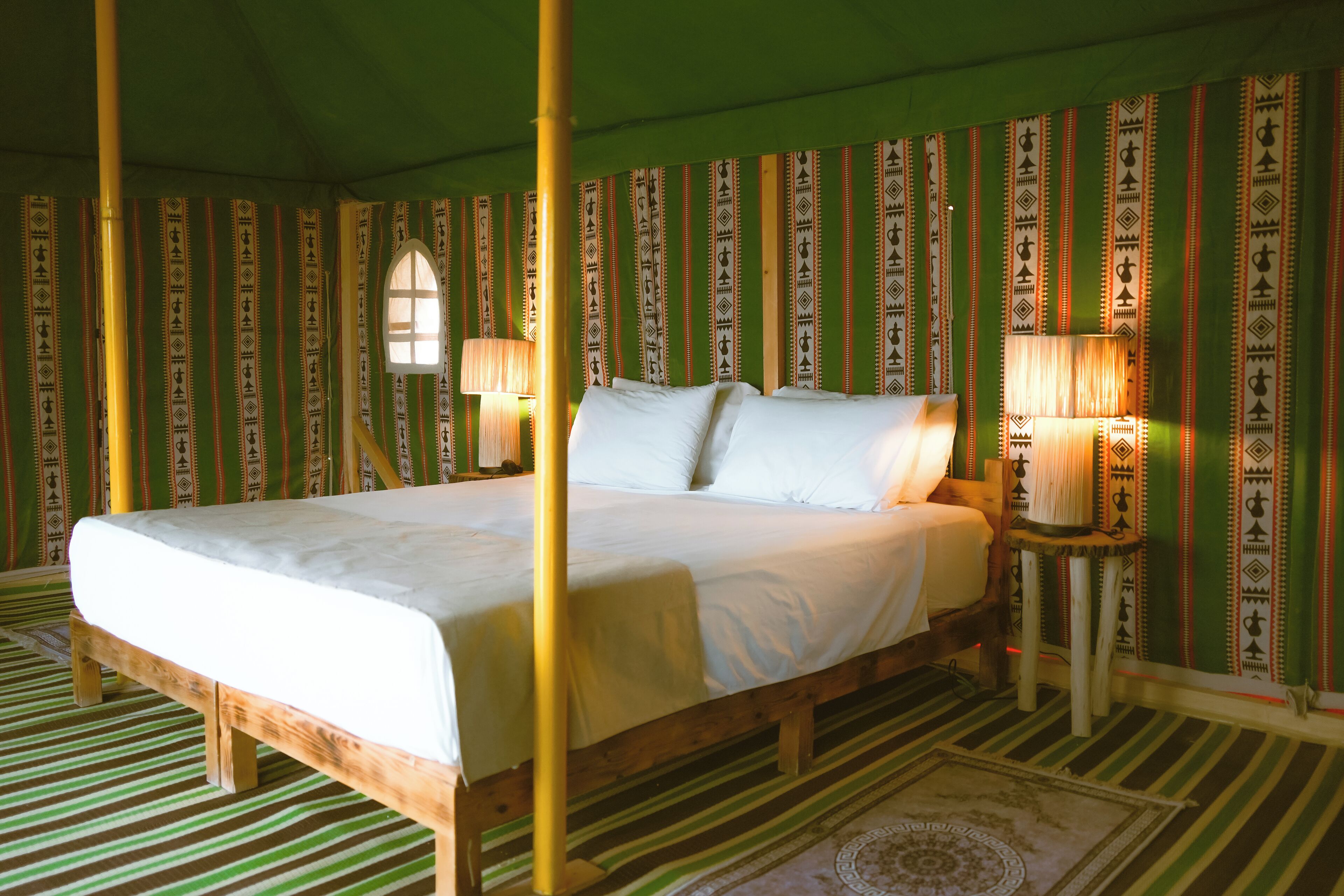 A room with a large bed, two bedside tables with lamps, a window, and patterned wallpaper.