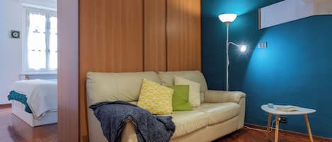 Een kamer met een beige bank, een houten garderobe, een blauwe muur, een vloerlamp en een kleine ronde tafel met een boek en een kop.