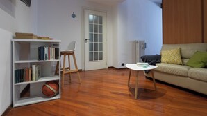 Un salon doté d’un plancher en bois, d’un canapé blanc avec des coussins, d’une table basse blanche, d’une bibliothèque et d’une porte avec des panneaux en verre dépoli.