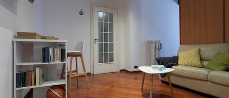 A living room with a wooden floor, a white sofa with cushions, a white coffee table, a bookshelf, and a door with frosted glass panels.
