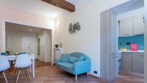 Un salon moderne avec un canapé bleu, un coin repas avec des chaises blanches et une cuisine aux carreaux de couleur turquoise.