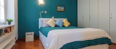 A bedroom with a bed, a bedside table, a nightstand, a window with curtains, a plant, and a wall-mounted light.